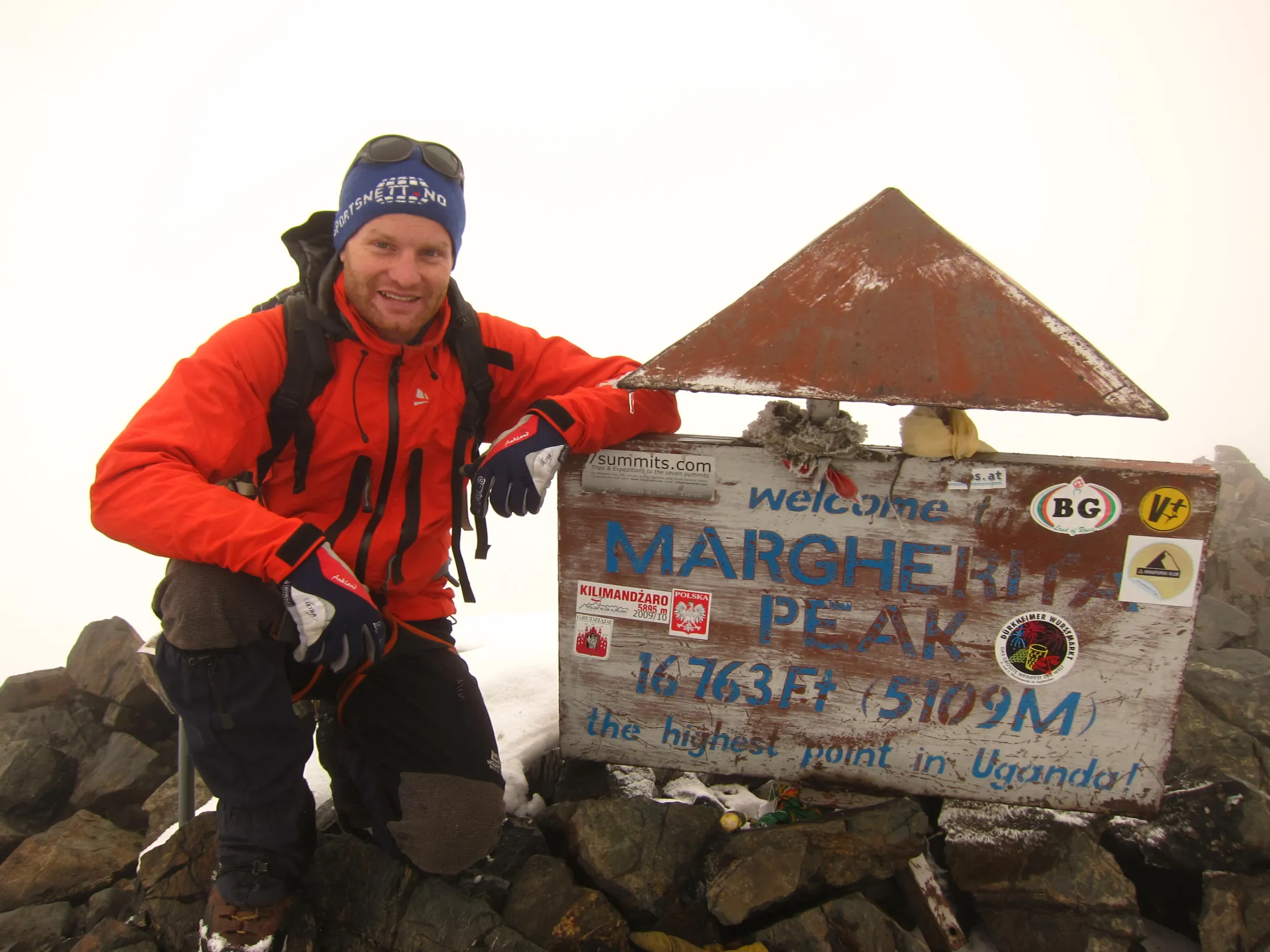 A smiling mountaineer in winter gear kneels on a rocky, snow-dusted summit next to a wooden sign marking Margherita Peak at 5109 meters.