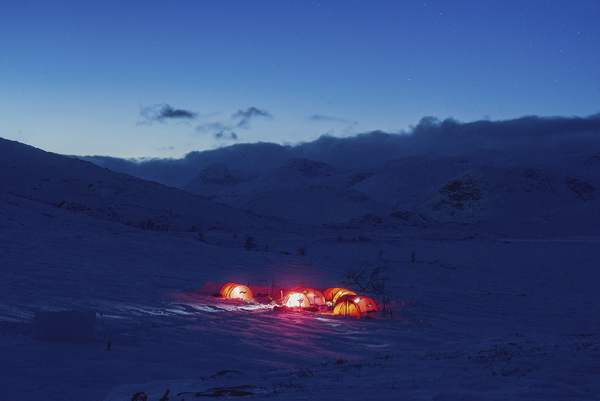 A distant view of glowing red tents pitched in a snow-covered mountain valley under a deep blue dusk sky.