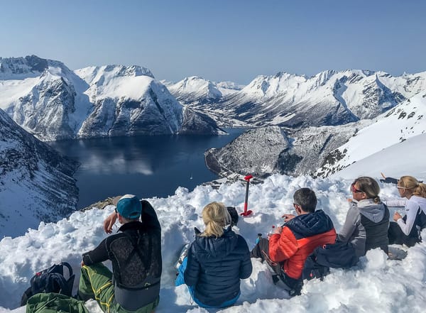 Five people sitting on a snow-covered mountain ridge, looking out over a fjord and snowy peaks.