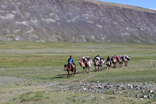 A caravan of pack camels loaded with gear is led by two riders on horseback across a grassy plain at the foot of a large, rocky mountain slope.
