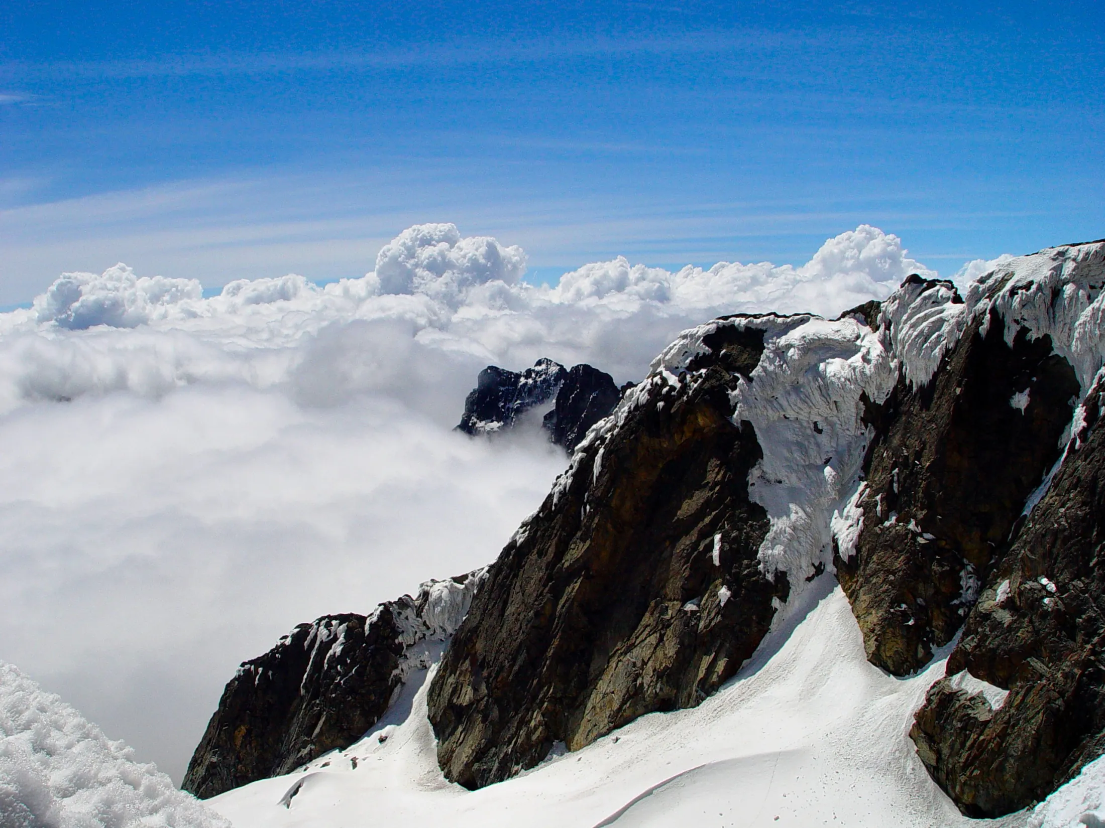 A dramatic high-altitude view of rugged, snow-covered mountain peaks rising sharply above a vast sea of thick white clouds under a clear blue sky.