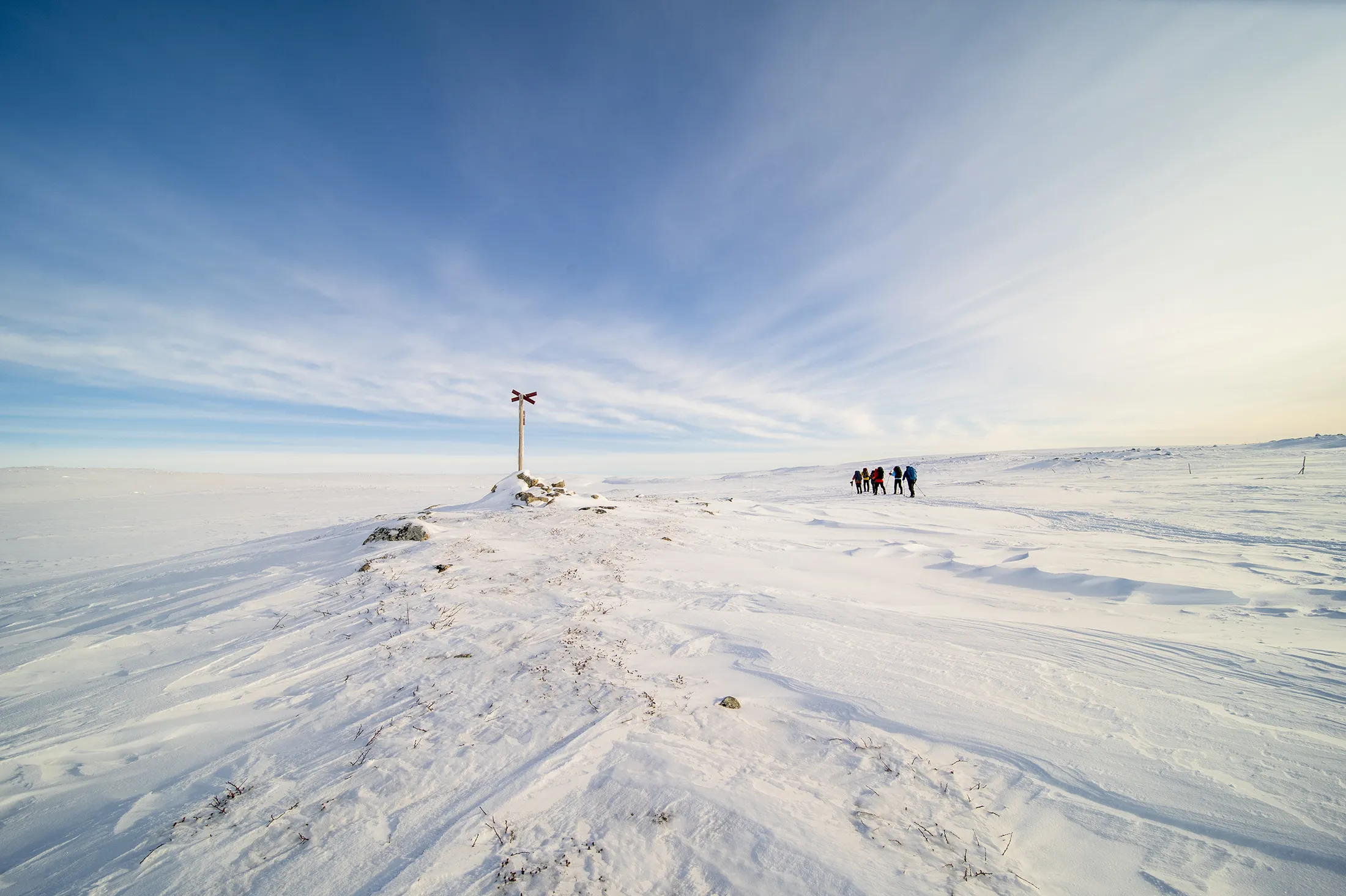 A snow-covered landscape with a solitary pole marker and a few distant figures under a bright blue sky.