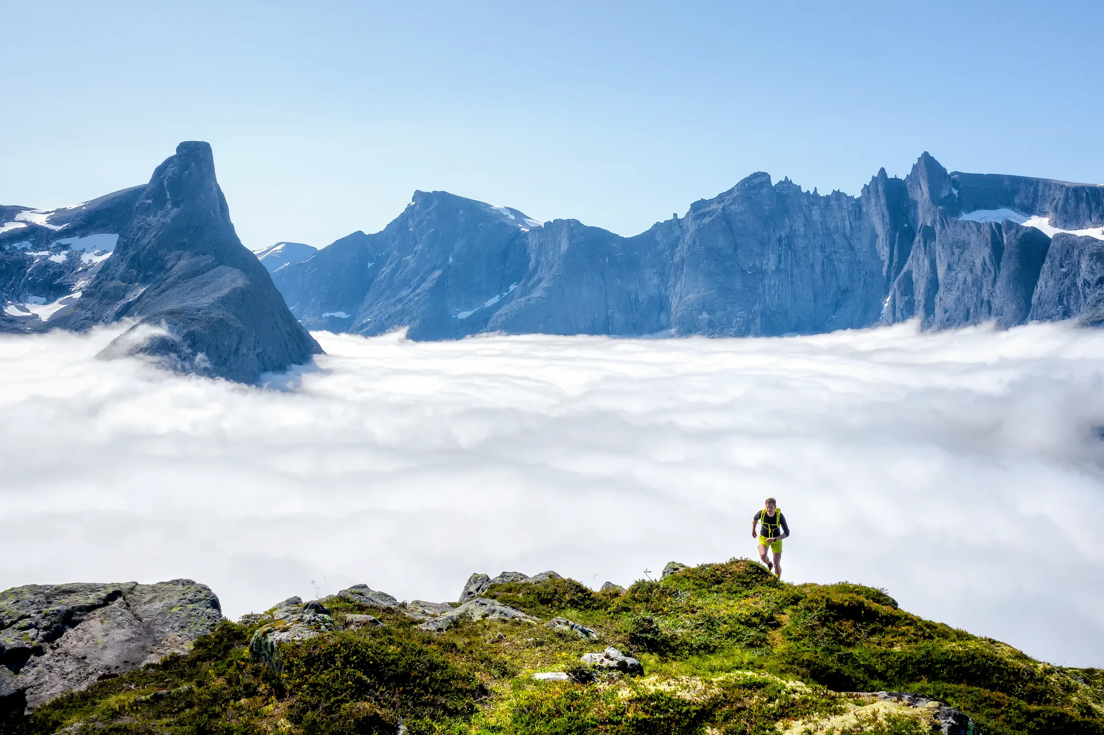 A hiker stands on a grassy mountain peak overlooking a sea of clouds with jagged peaks in the distance.