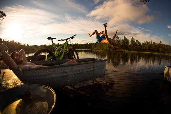 A man relaxing in a metal tub beside a lake while another man dives into the water.