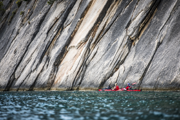 A low-angle shot shows two people in a red tandem sea kayak paddling close to the base of a massive, steeply angled, textured rock cliff that rises directly out of the deep blue water.