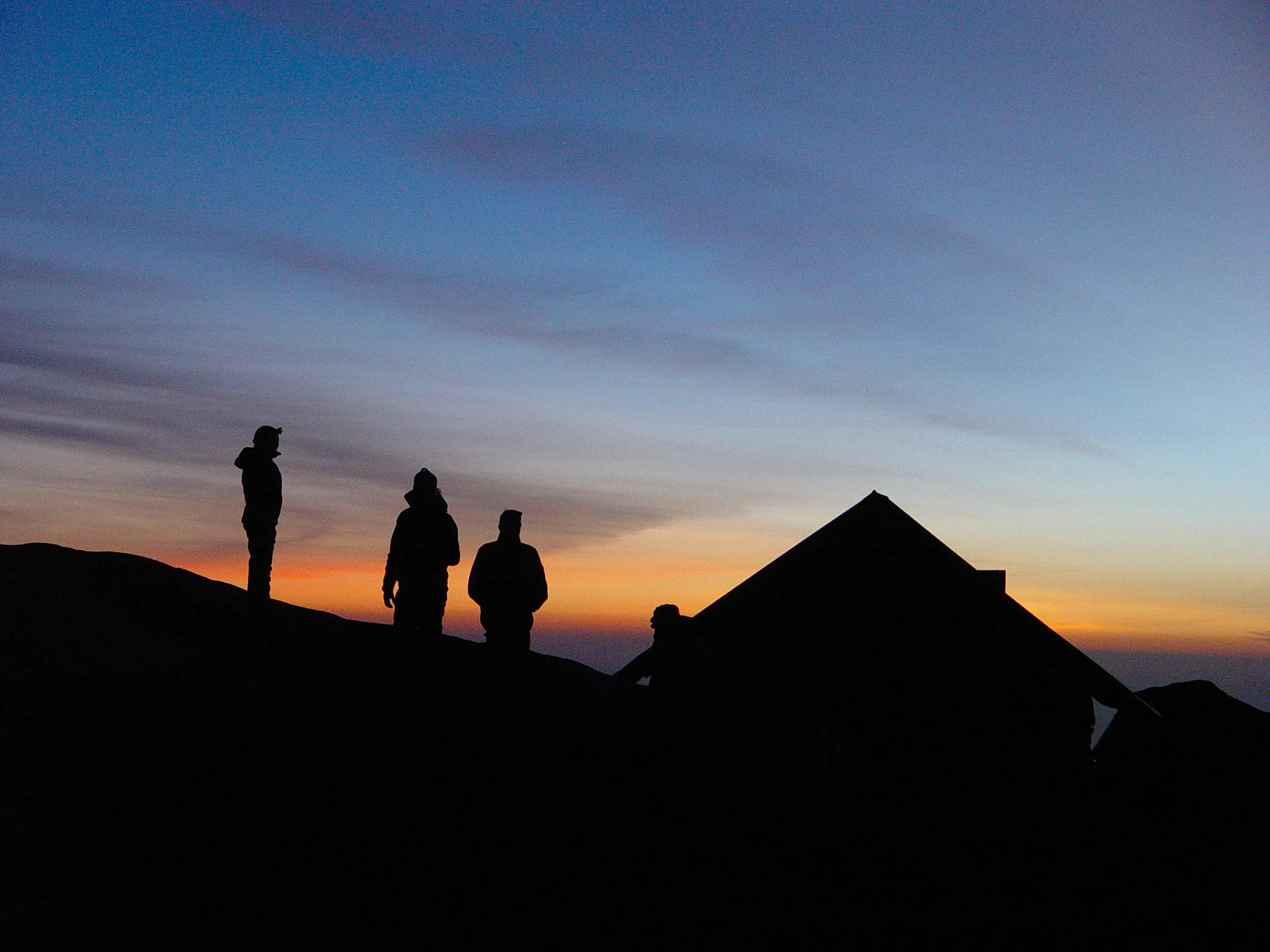 The silhouettes of several people and a small mountain hut stand out against a dramatic sunset sky with vibrant shades of blue, orange, and yellow.