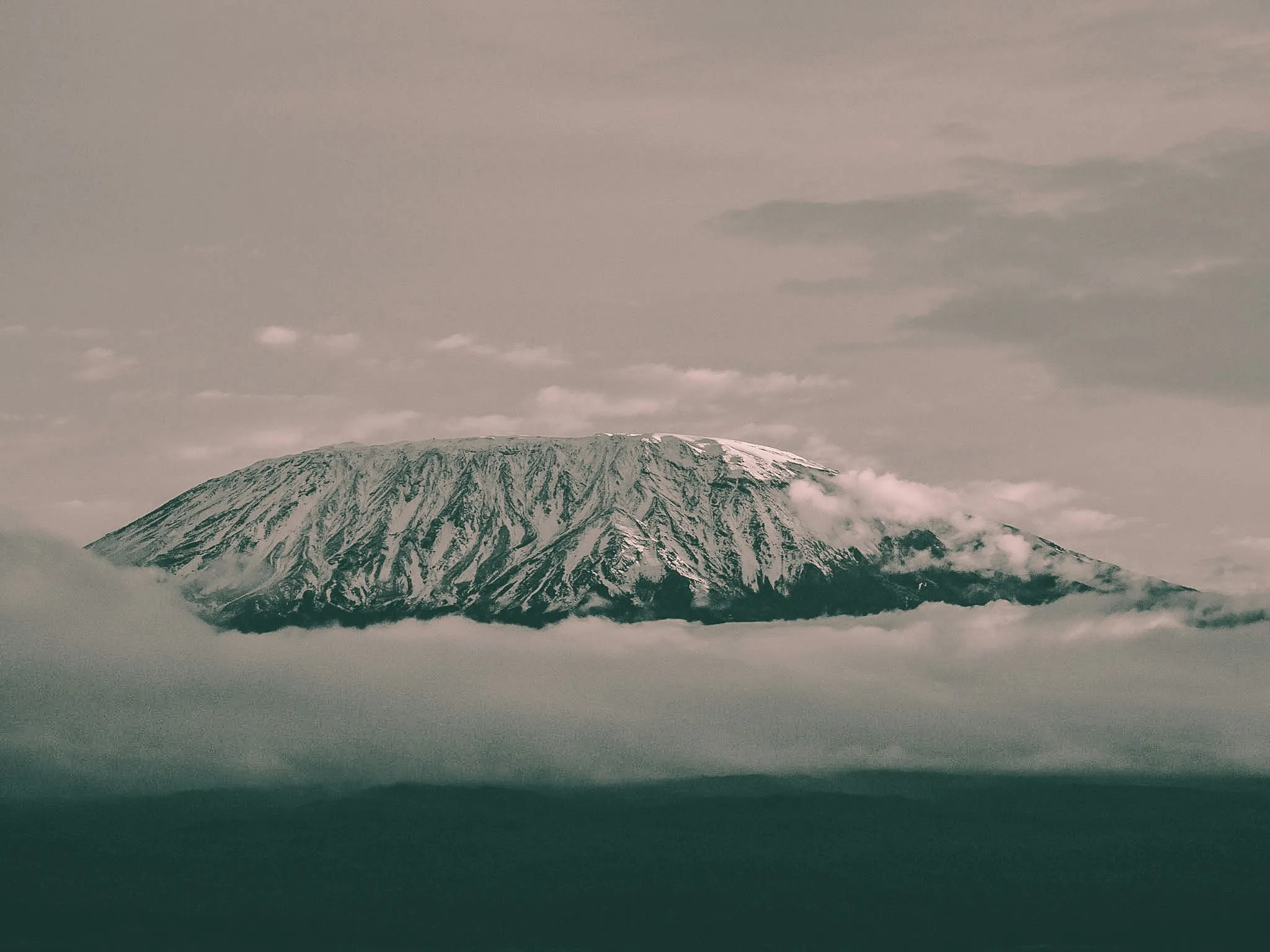 The snow-covered, rocky peak of Mount Kilimanjaro looms above a thick bed of clouds under an overcast sky.