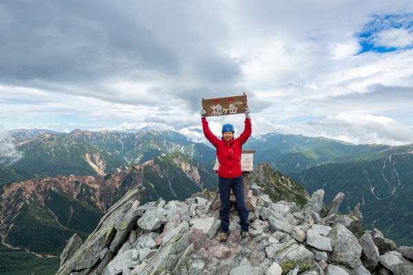 A smiling hiker wearing a red jacket and blue helmet stands on a rocky summit, holding up a wooden sign with Japanese text displaying an elevation of 3180m, with green mountains stretching into the distance.