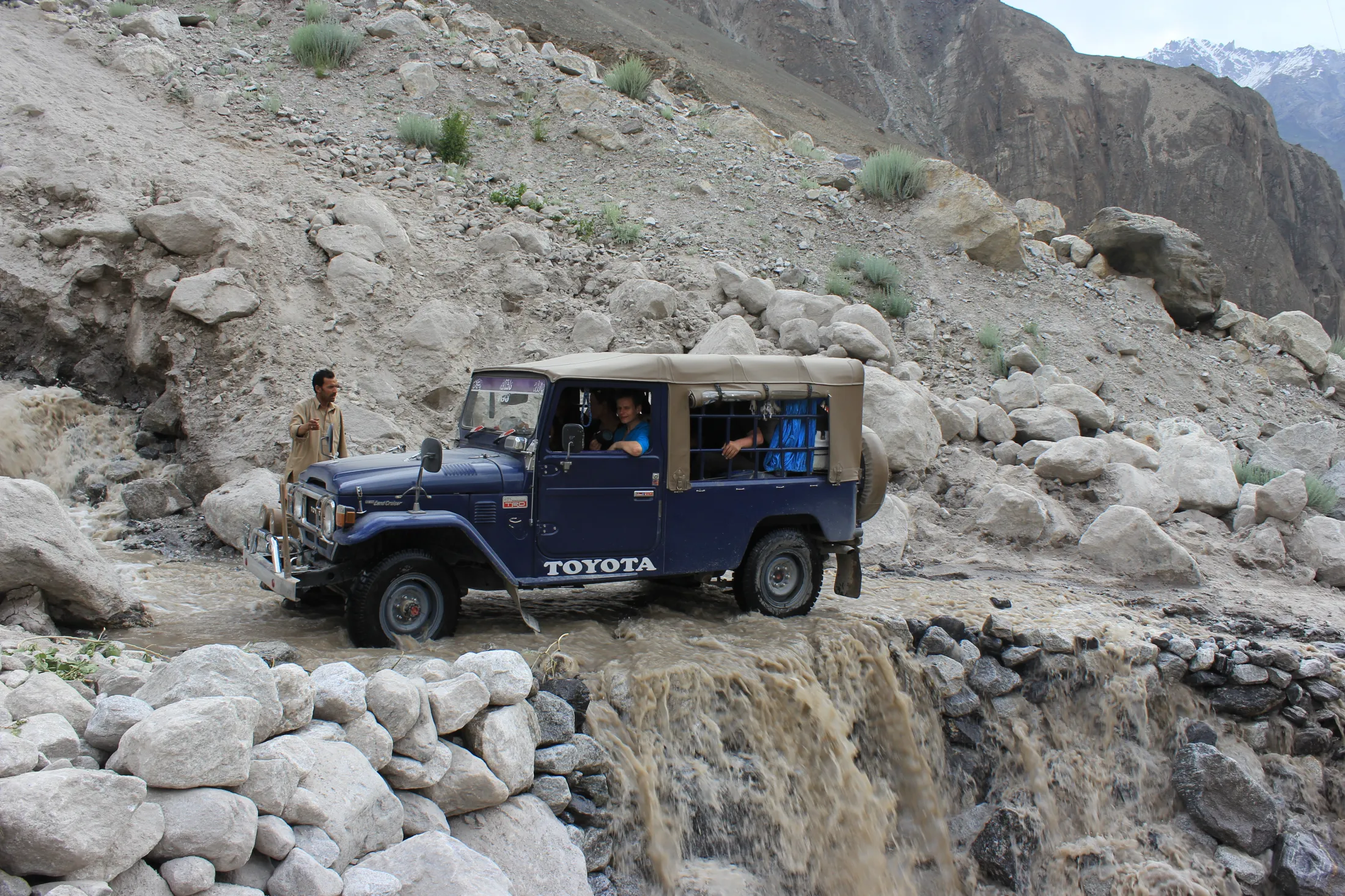 A blue Toyota 4x4 vehicle crosses a rushing, muddy river stream along a rugged, rocky mountain road.