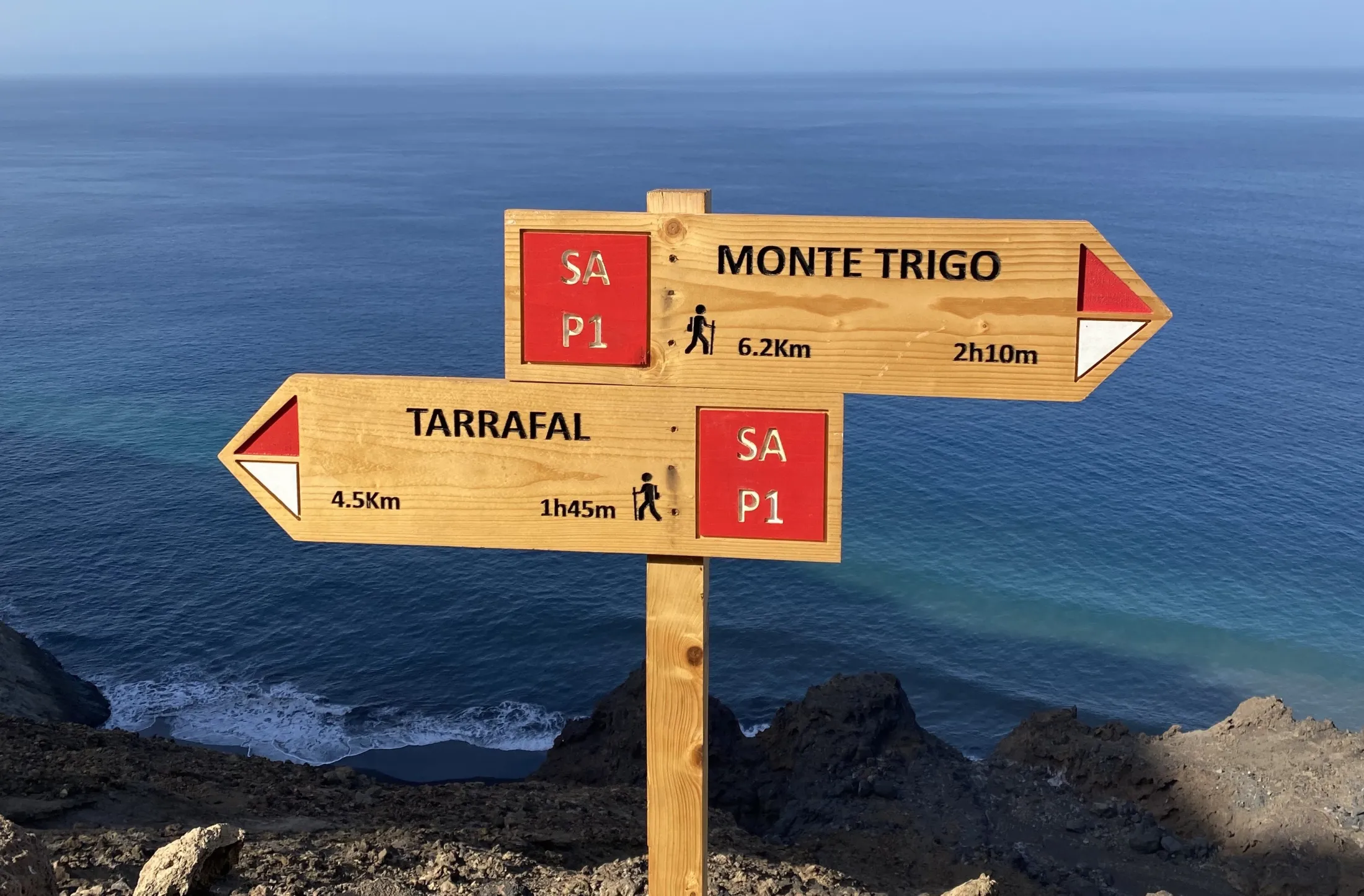A wooden hiking signpost stands on a cliffside, pointing left to Tarrafal and right to Monte Trigo, set against a vast backdrop of the blue ocean.