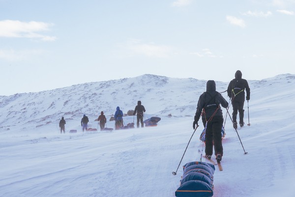 A line of skiers pulling pulks up a snowy, windswept mountain slope.