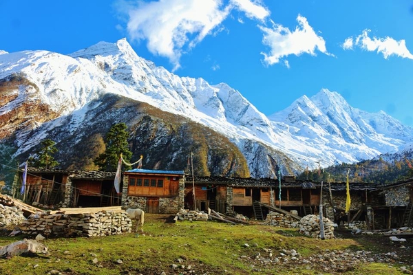 Traditional wooden and stone buildings sit on a grassy slope, backlit by towering, snow-covered mountain peaks under a bright blue sky with scattered clouds.