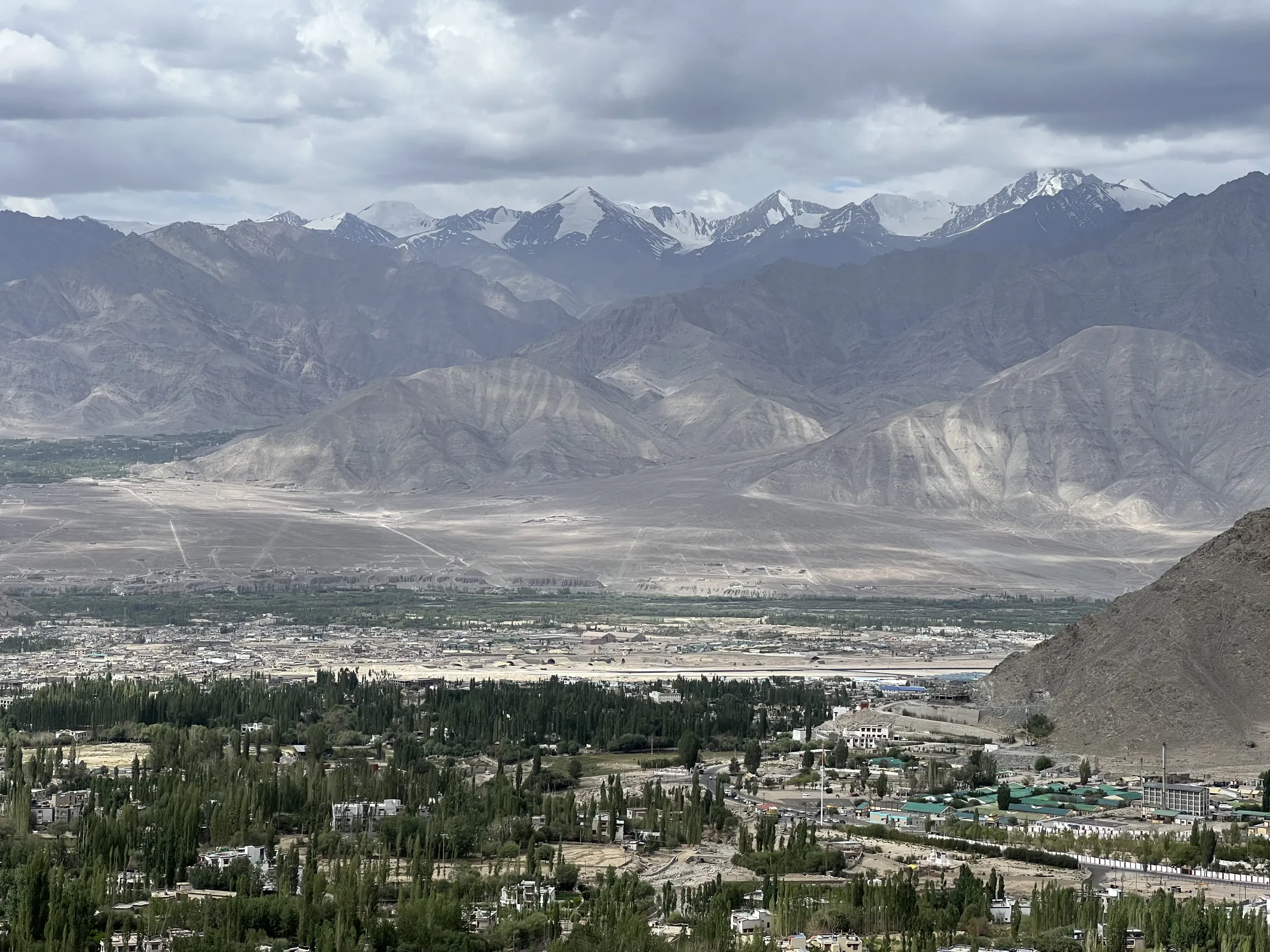 A high-altitude valley town with lush green trees sits in the foreground, contrasted by barren foothills and a dramatic backdrop of snow-capped mountain peaks under an overcast sky.