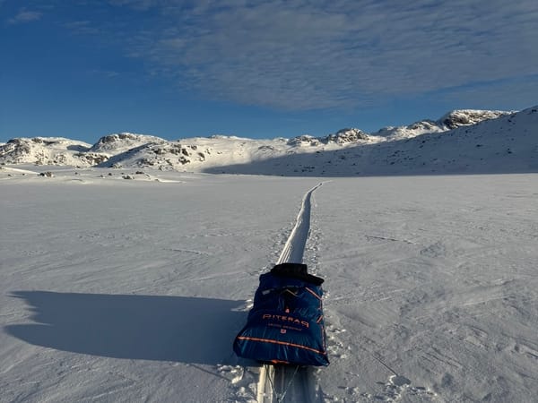 A pulk sits on cross-country ski tracks in a vast, snow-covered mountain landscape under a blue sky.