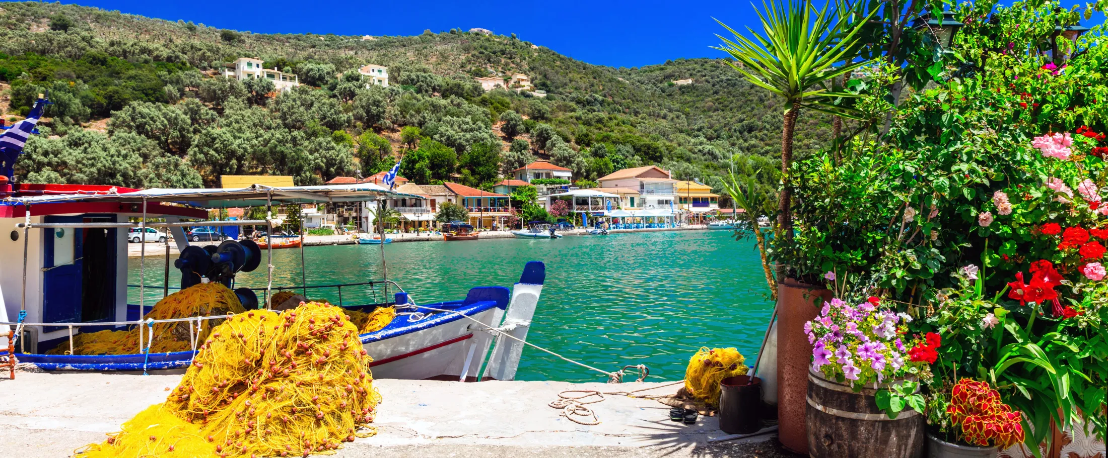 A vibrant harbor scene featuring a traditional blue and white fishing boat tied to a concrete pier piled with bright yellow fishing nets, looking across calm turquoise water toward a coastal village nestled on a lush, green hillside under a clear blue sky.