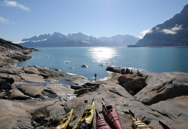 Kayaks are pulled up onto the rocky shore in the foreground while a group of people rests, and one person bathes in the sun-drenched ocean surrounded by majestic mountains.