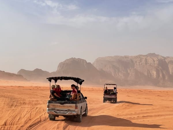 Two open pickup trucks carrying tourists drive across orange desert sands toward distant mountain formations.