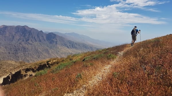A hiker walks along a dirt trail on a golden, grassy mountain slope that overlooks a vast, rugged valley stretching toward a distant mountain range under a blue sky.
