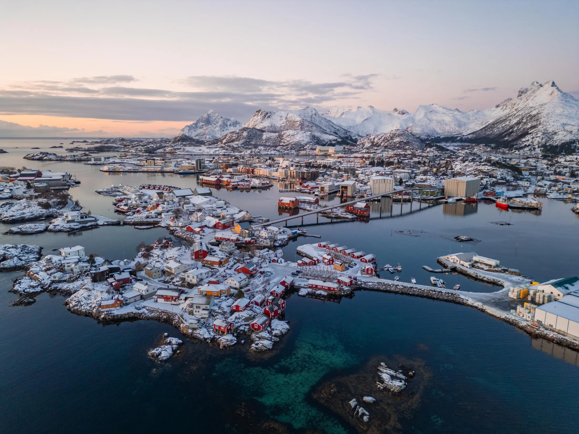 Drone photo of Svolvær new, with Svinøya rorbuer (fisherman’s cabins) in the foreground on an island. Red cabins, white mountains, and a snow-covered town, deep blue ocean, and a bright sky