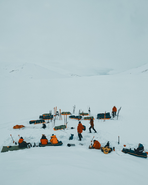 A group of skiers taking a break on the snow, sitting and resting near their equipment and sleds in a vast arctic landscape.