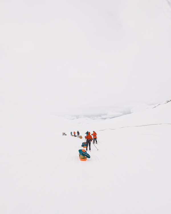 Skiers in orange jackets pull expedition sleds single file across a vast, white arctic landscape under a pale sky.