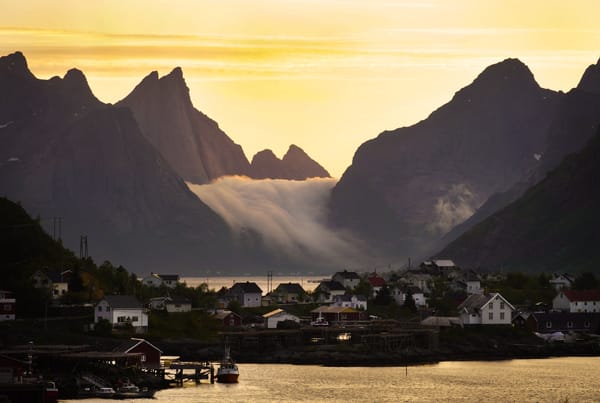 A coastal town nestled beneath steep mountains in Lofoten, Norway, with low clouds spilling between the peaks under a golden sunset.