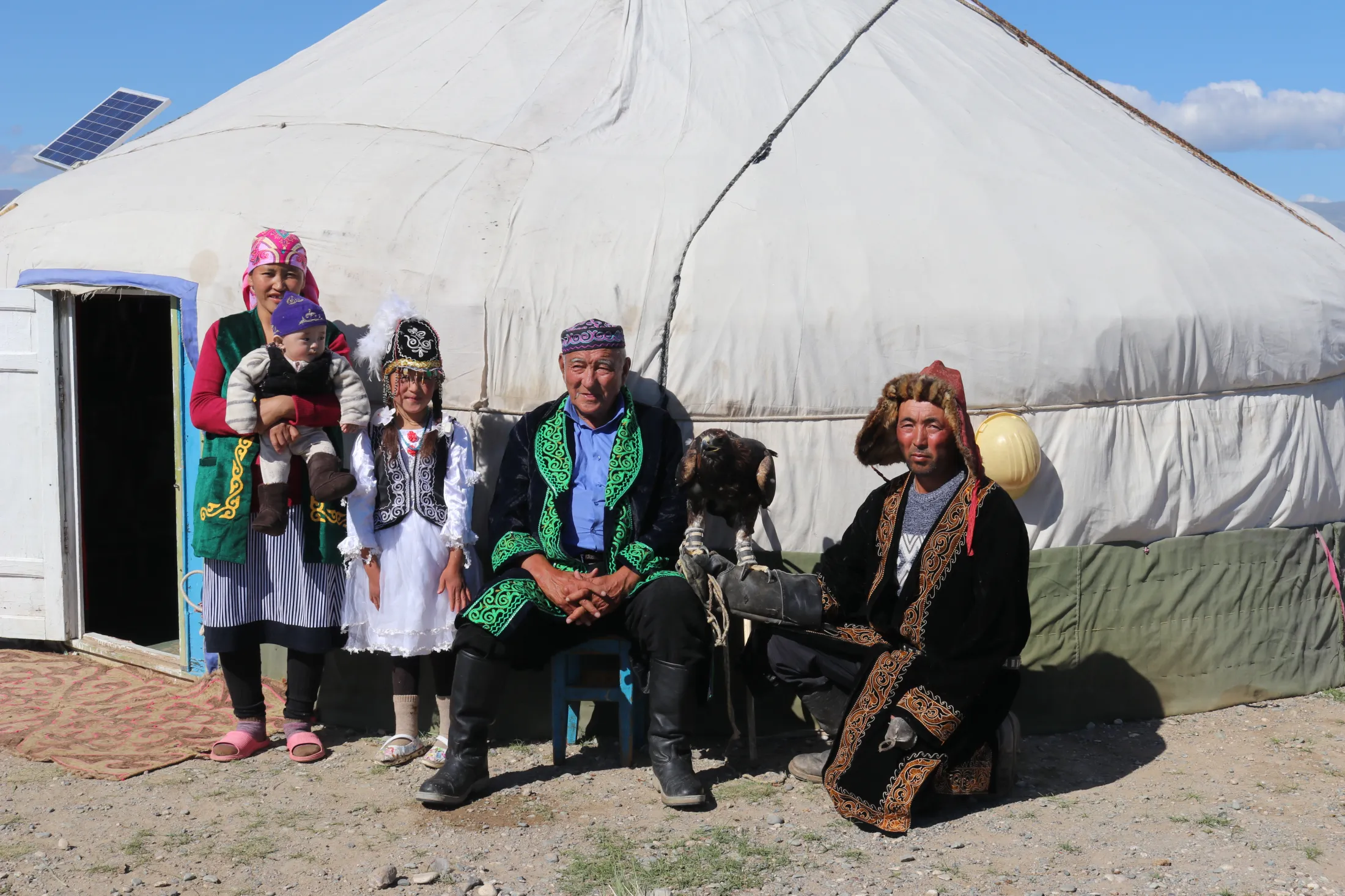 A family wearing ornate traditional attire poses outside a large white yurt, including a man holding a perched golden eagle on his gloved hand.