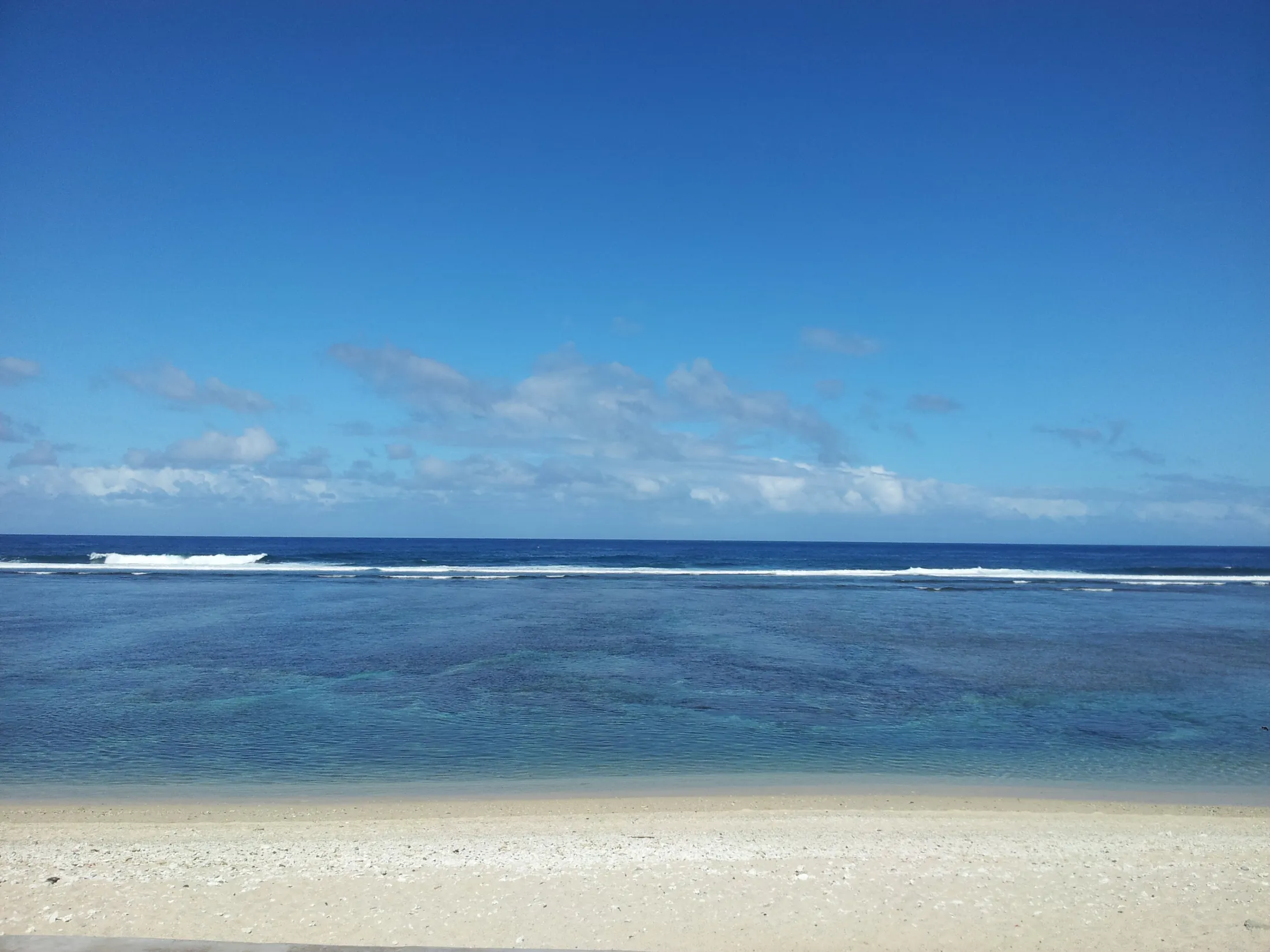 A scenic view of a sandy beach looking out over calm, turquoise lagoon waters toward breaking white waves on a distant coral reef under a blue sky.