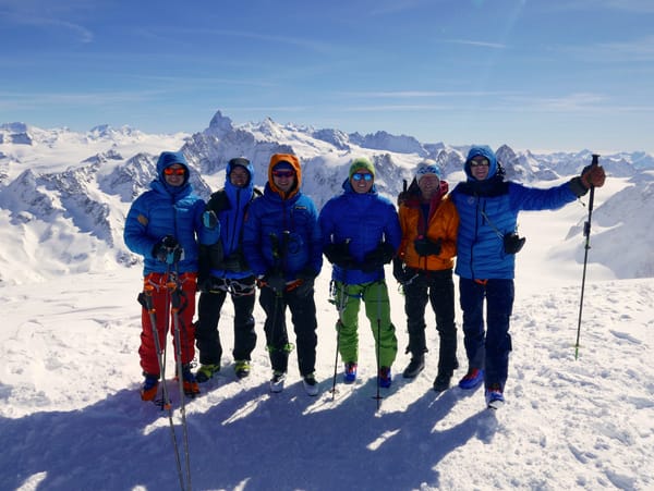 A group of six mountaineers in colorful winter gear and sunglasses pose together on a snowy peak with a vast panorama of Alpine mountains behind them.