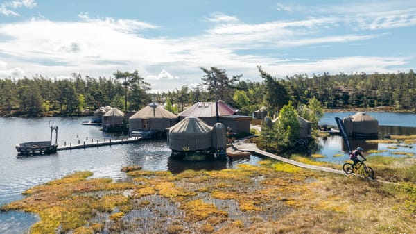 A collection of yurts and wooden buildings situated on a lake with a person on a bicycle on a wooden path.