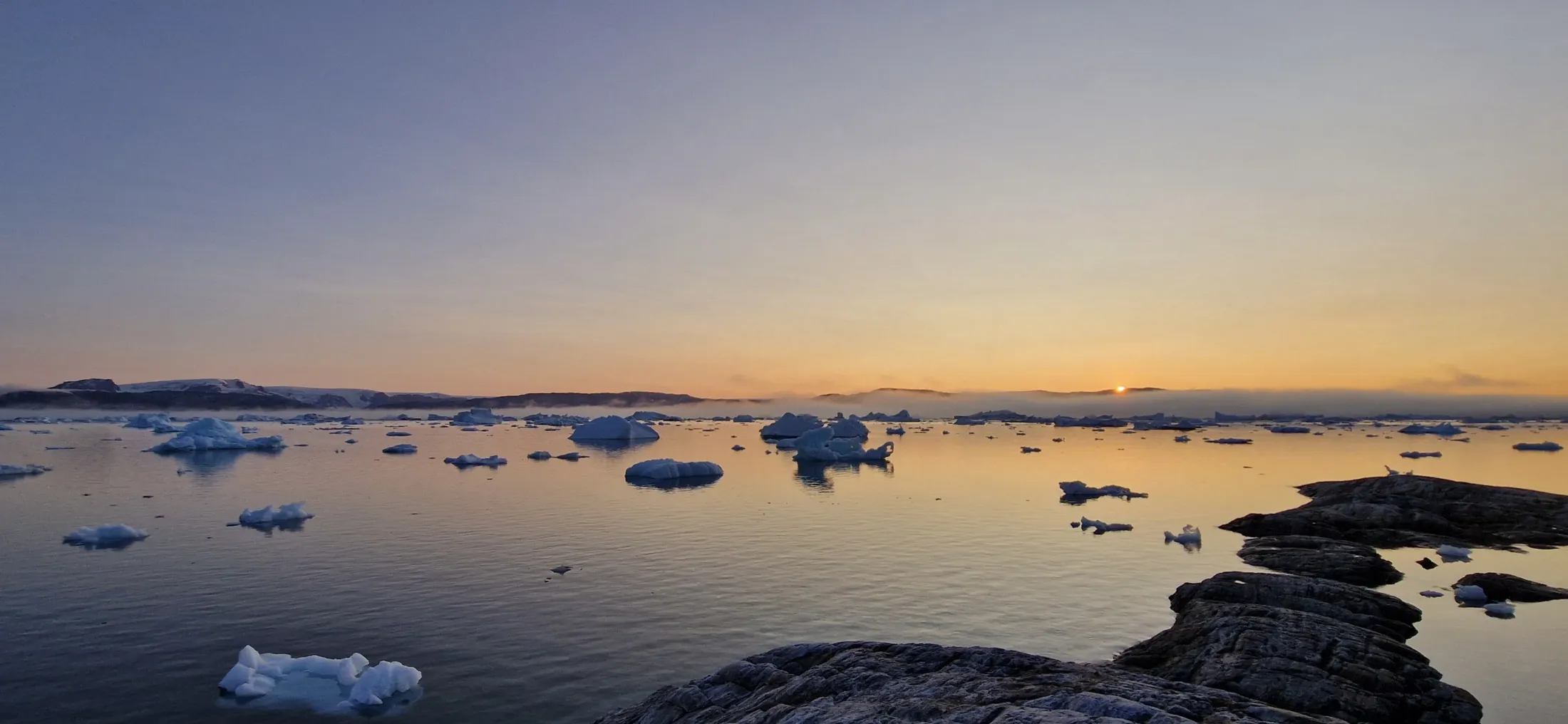 Icebergs float on a calm ocean during a golden sunset, with low fog hanging over mountains in the background and rocky terrain in the foreground.
