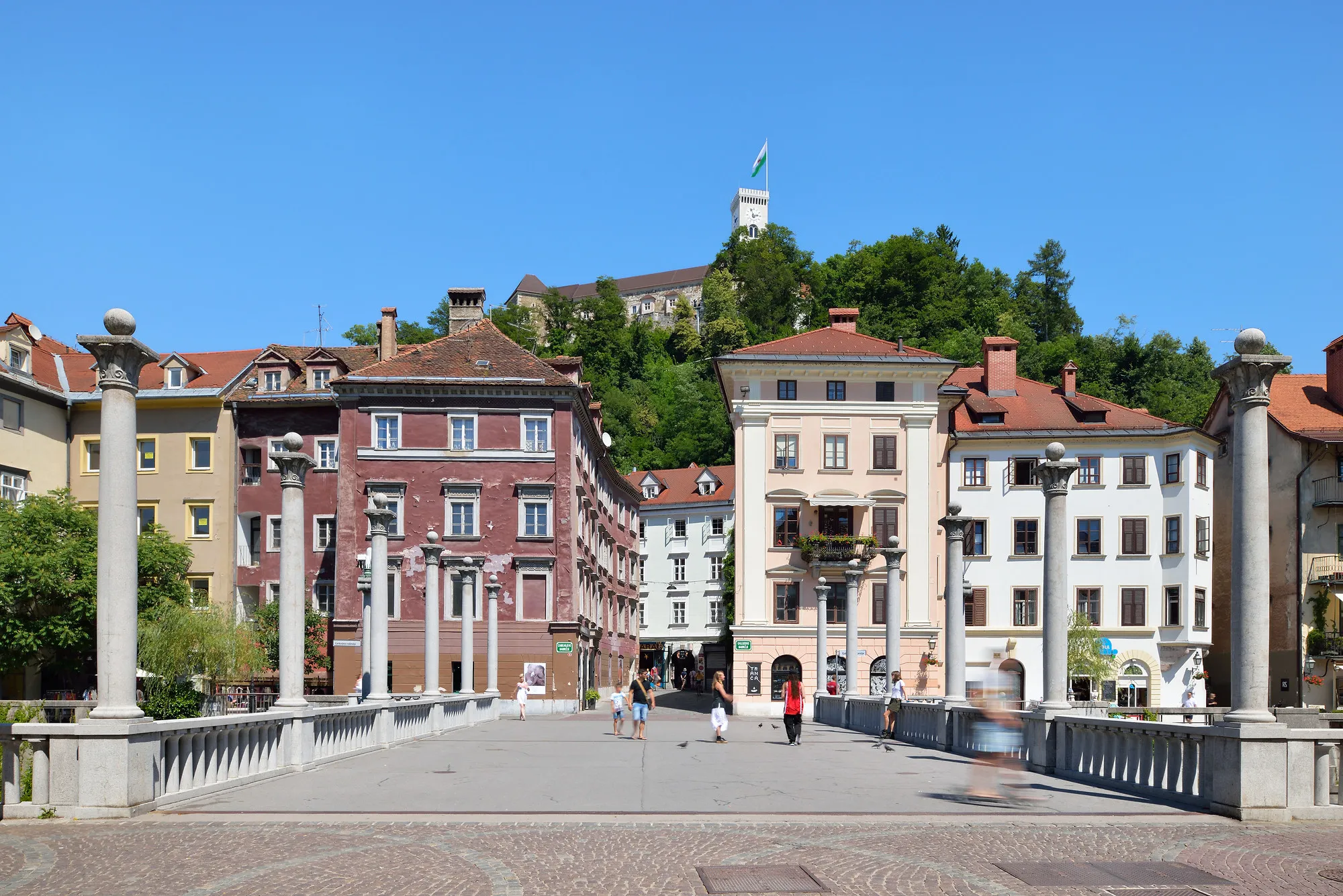 Pedestrians walk across a wide, stone-pillared bridge leading toward colorful historic buildings, with Ljubljana Castle standing prominently on a green hill under a clear blue sky.