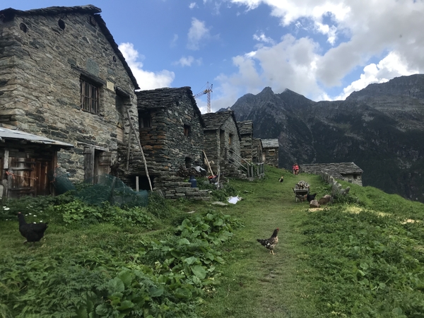 A row of traditional stone houses sits on a grassy slope in an alpine village, with chickens wandering the path and rugged mountain peaks rising in the background under a cloudy sky.