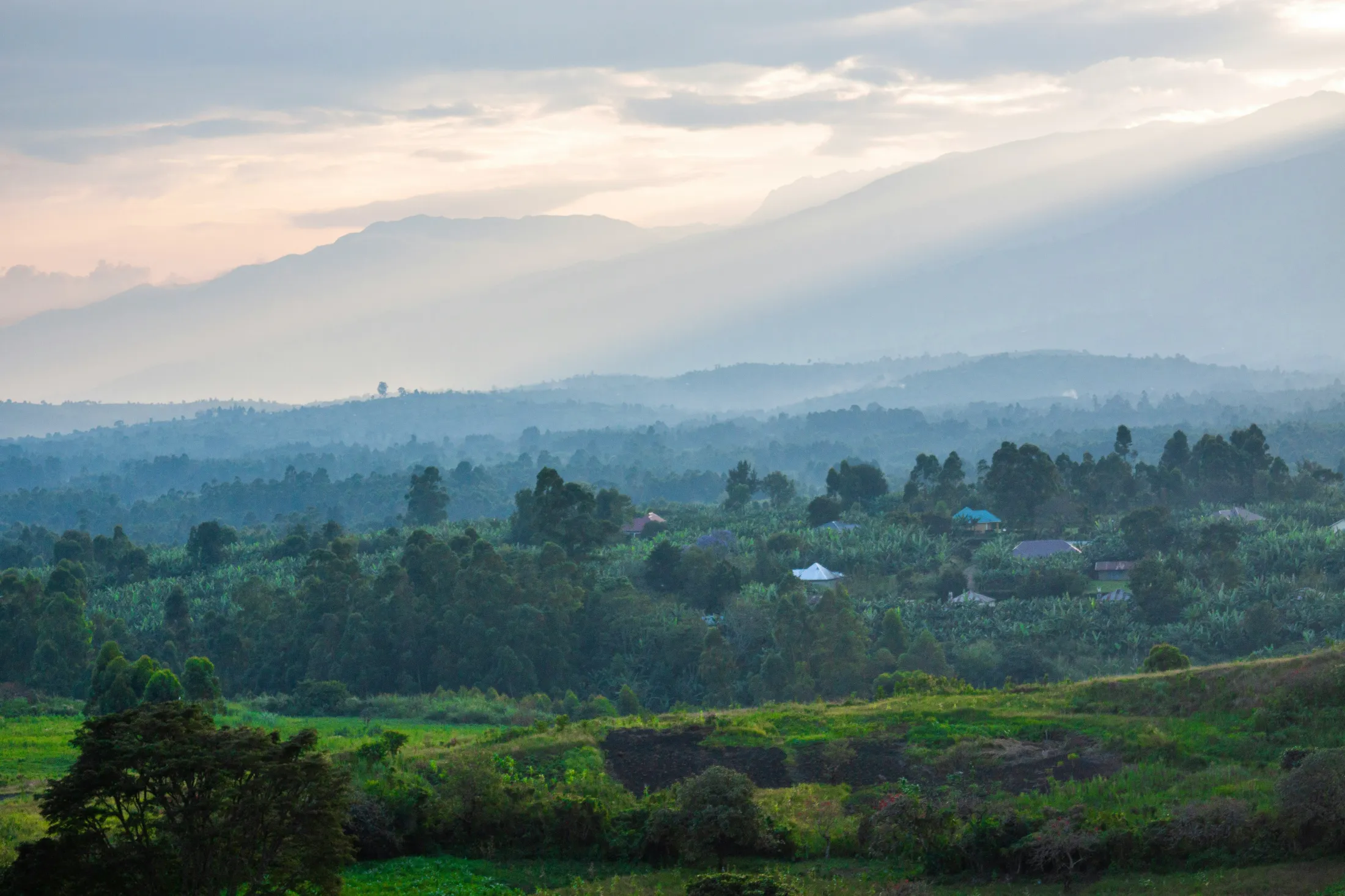Sunlight filters through clouds over a lush, hazy green landscape with rolling hills, scattered houses, and distant layers of mountains.