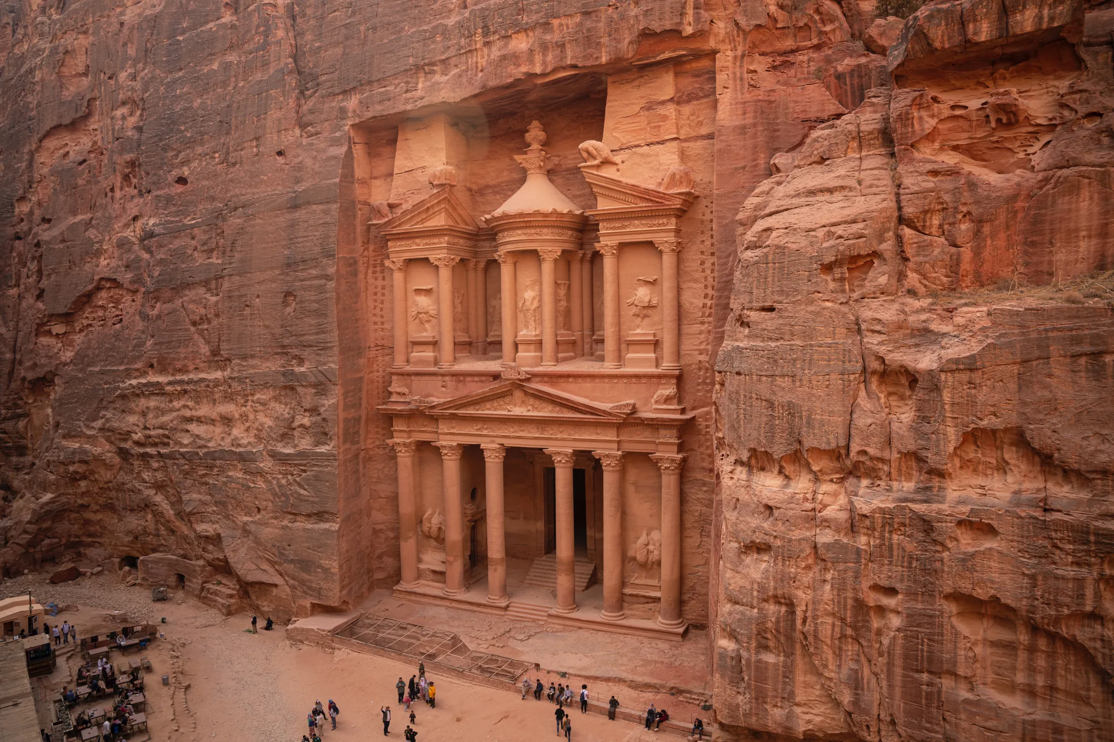 The carved facade of The Treasury (Al-Khazneh) in Petra sculpted directly into the reddish-brown sandstone cliff, with small tourists visible on the ground below.