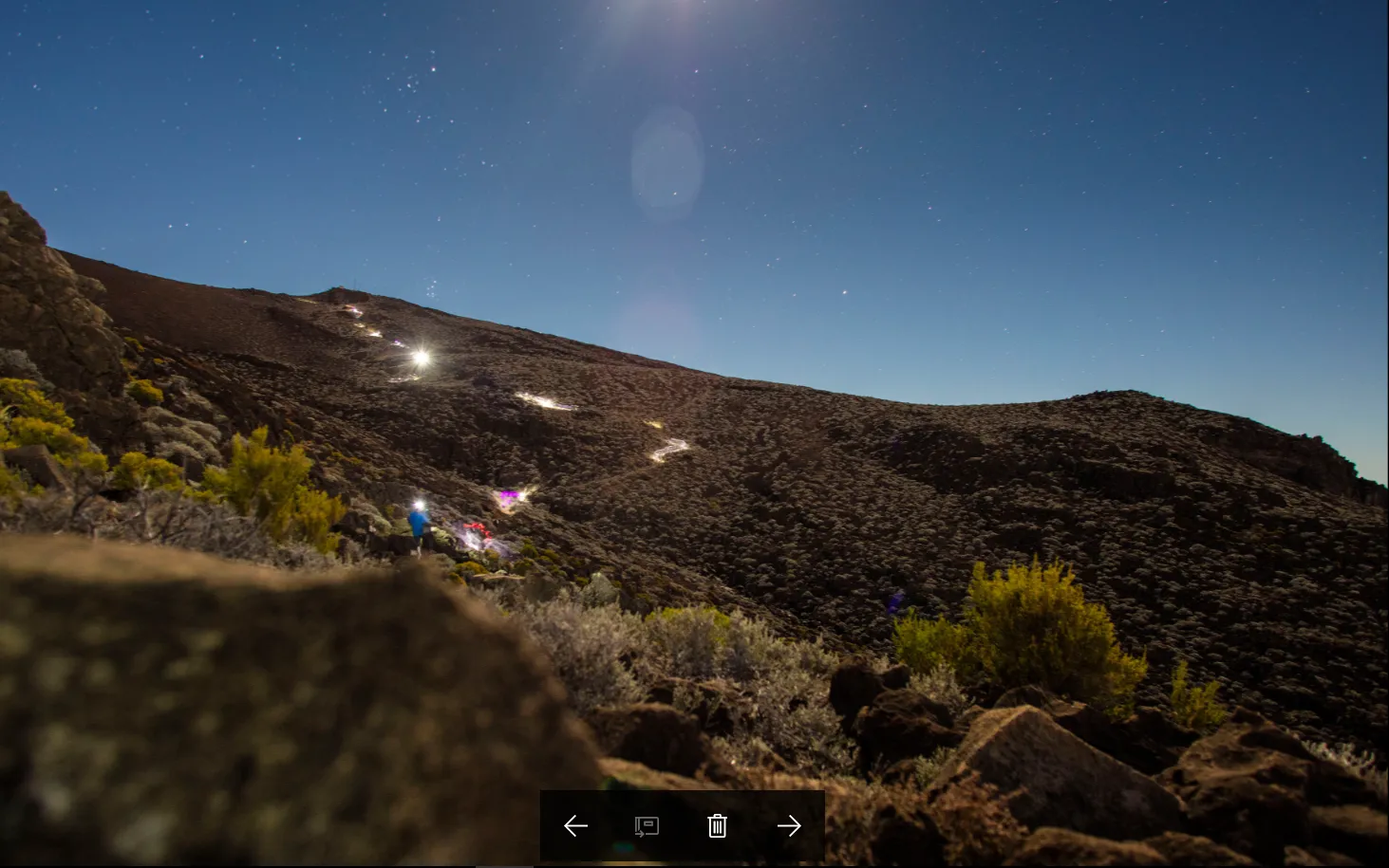 A winding line of hikers' headlamps illuminates a steep mountain trail under a clear, starry night sky.
