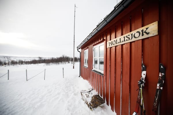 A red wooden cabin named 'Mollisjok' in a snowy, desolate landscape, with skis leaning against the wall.
