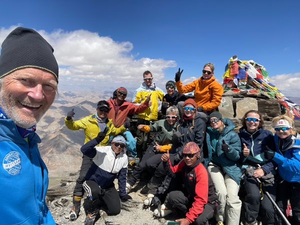 A smiling group of mountaineers poses together at a rocky summit under a clear blue sky, next to a pile of stones covered in colorful prayer flags.