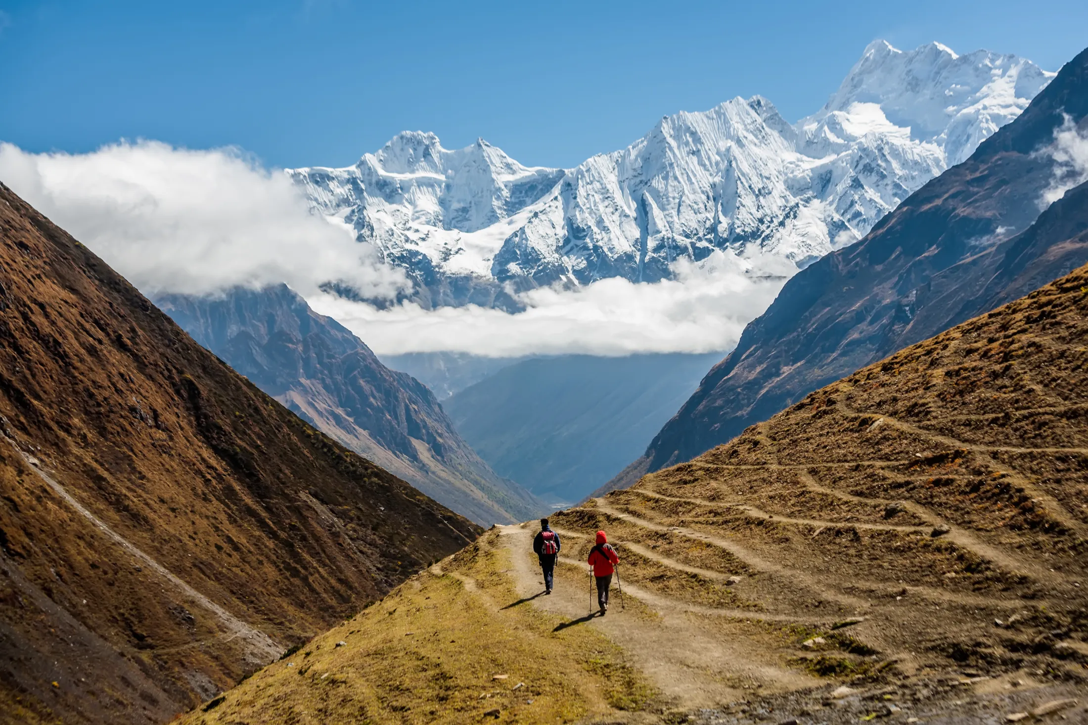 Two hikers walk down a dirt path in a vast mountain valley, heading toward towering, snow-capped peaks partially covered by clouds under a clear blue sky.