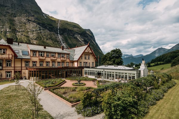 A large traditional hotel with a garden and a greenhouse, set against dramatic mountains in a lush valley.