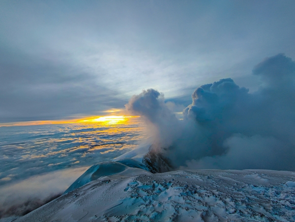 A snow-covered mountain peak with a plume of smoke or steam rising from it at sunrise.