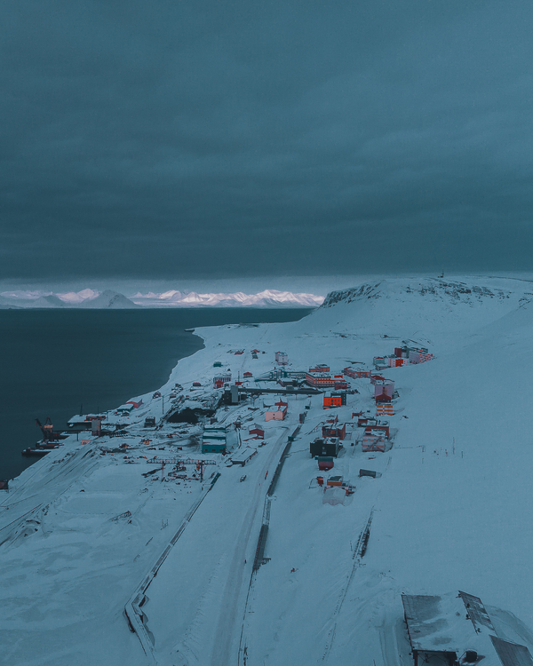 An aerial view of a snow-covered coastal settlement next to dark arctic waters, with mountains lit by distant sunlight under a heavy, blue-grey sky.