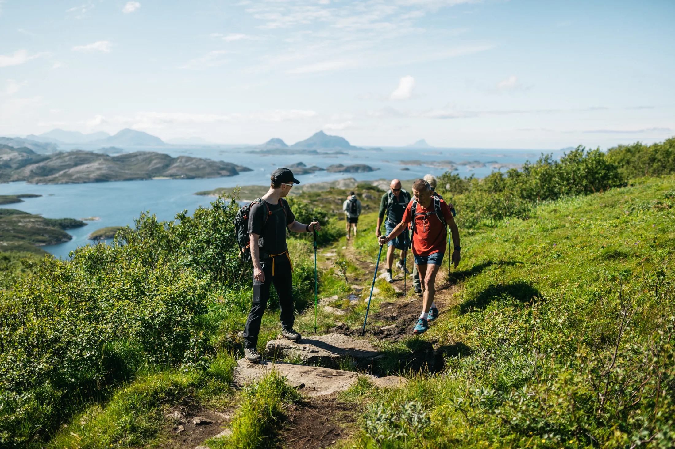 Hikers walking on a scenic coastal trail in Helgeland, Norway, with ocean and islands in the background.
