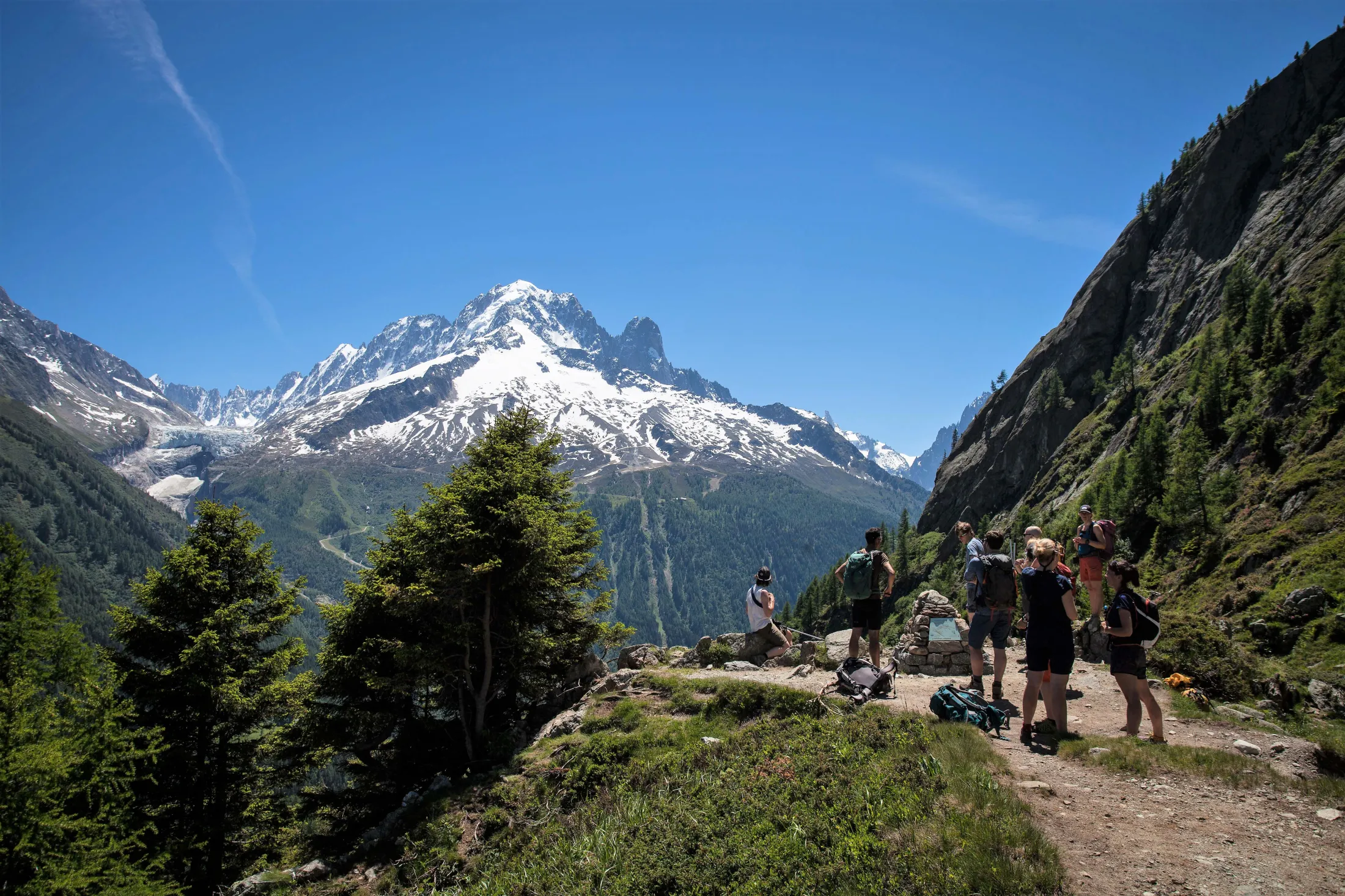 People on a trail in mountains looking at a larger mountain, blue skies.
