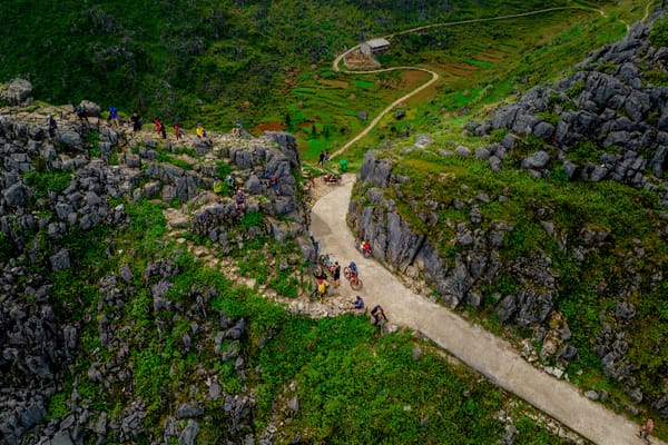 An aerial view of a group of hikers walking along a dirt trail winding through a rocky, green landscape.
