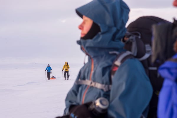 A person in a blue hooded jacket standing in a snowy landscape, with other skiers in the distance.