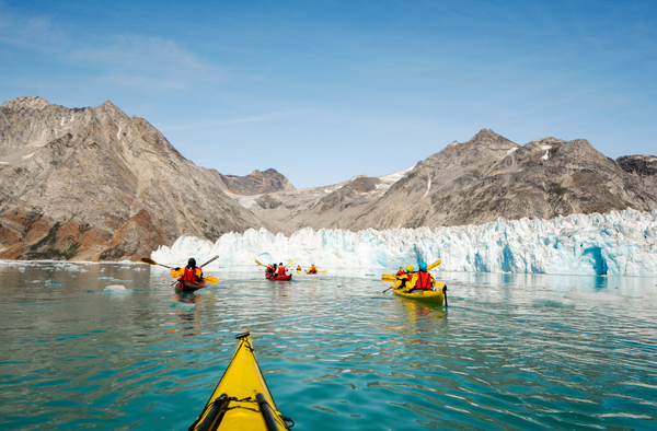 View from a yellow kayak toward a group of kayaks paddling on light green water toward a massive glacier wall bordered by tall, barren mountains under a clear sky.
