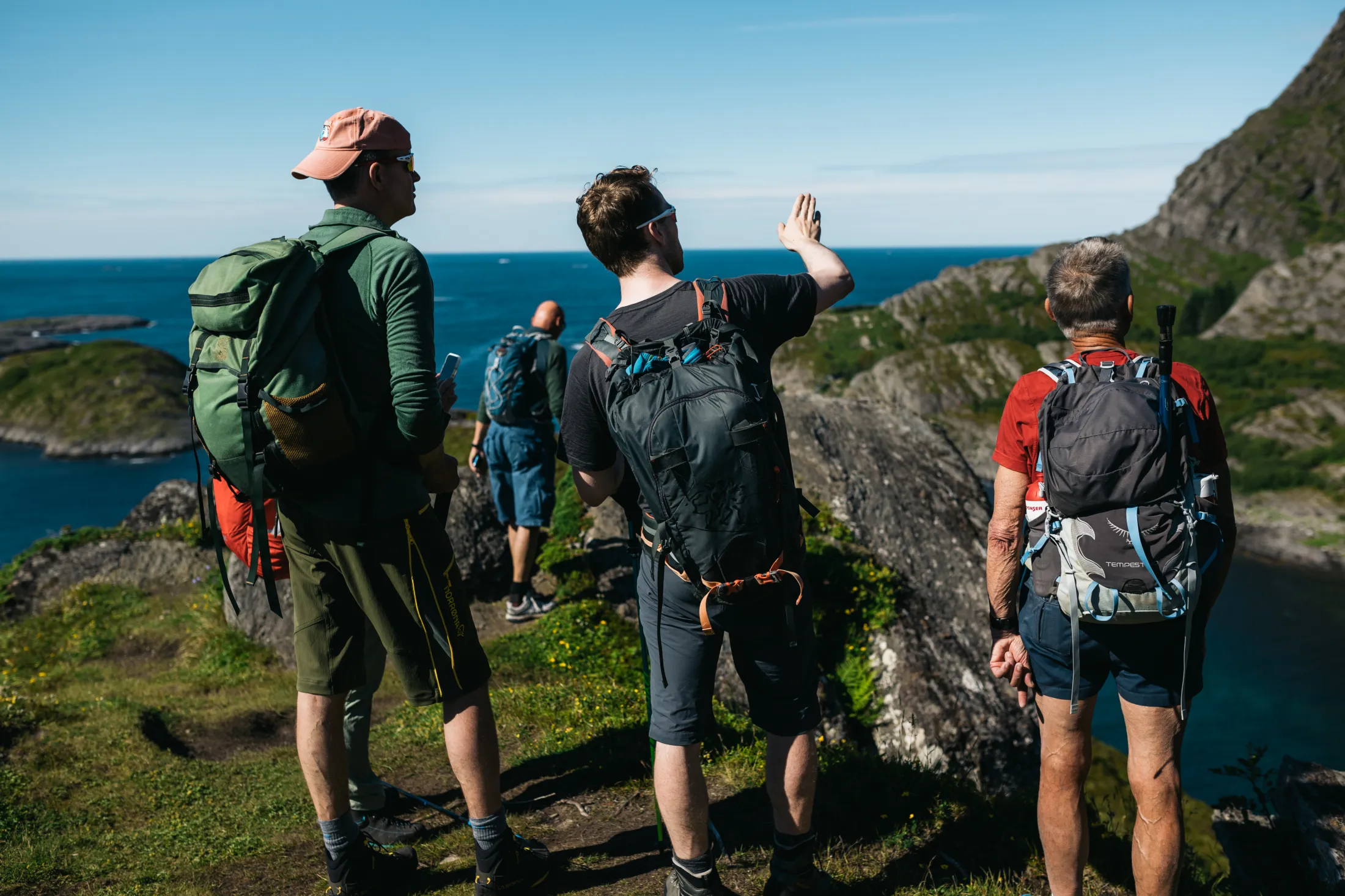 Four hikers with backpacks look out toward the ocean from a cliff top, with one pointing in the distance.