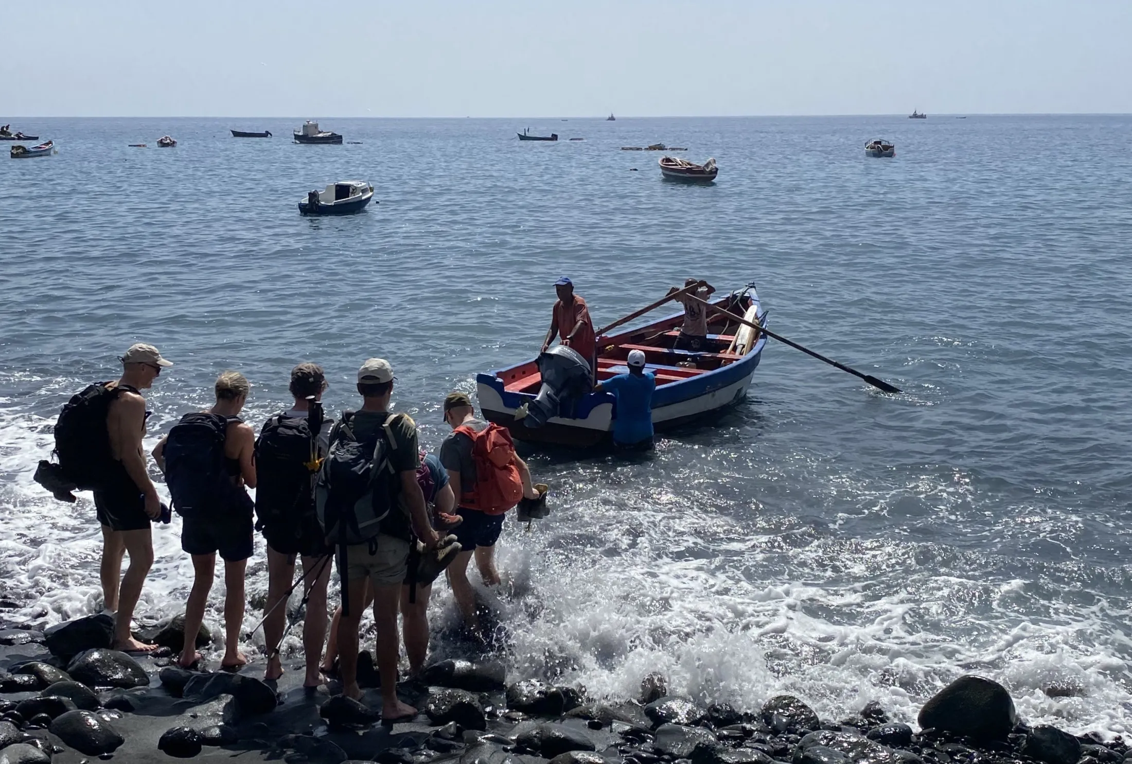 A group of hikers carrying backpacks wade through the surf on a rocky beach toward a small wooden boat manned by locals, with several other boats scattered across the calm ocean behind them.