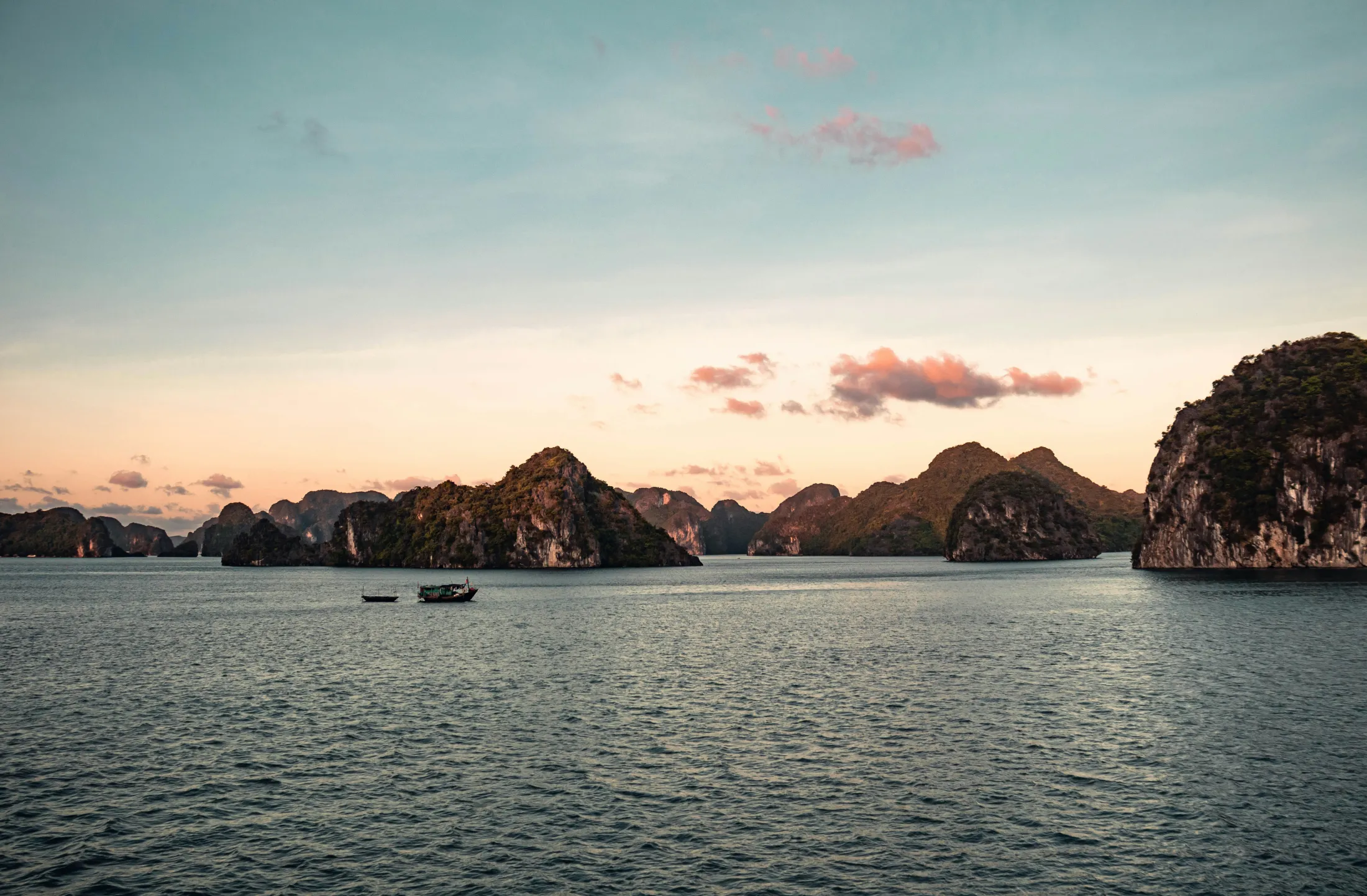 Rugged limestone islands rising from calm waters during a colorful sunset, with small boats floating nearby.